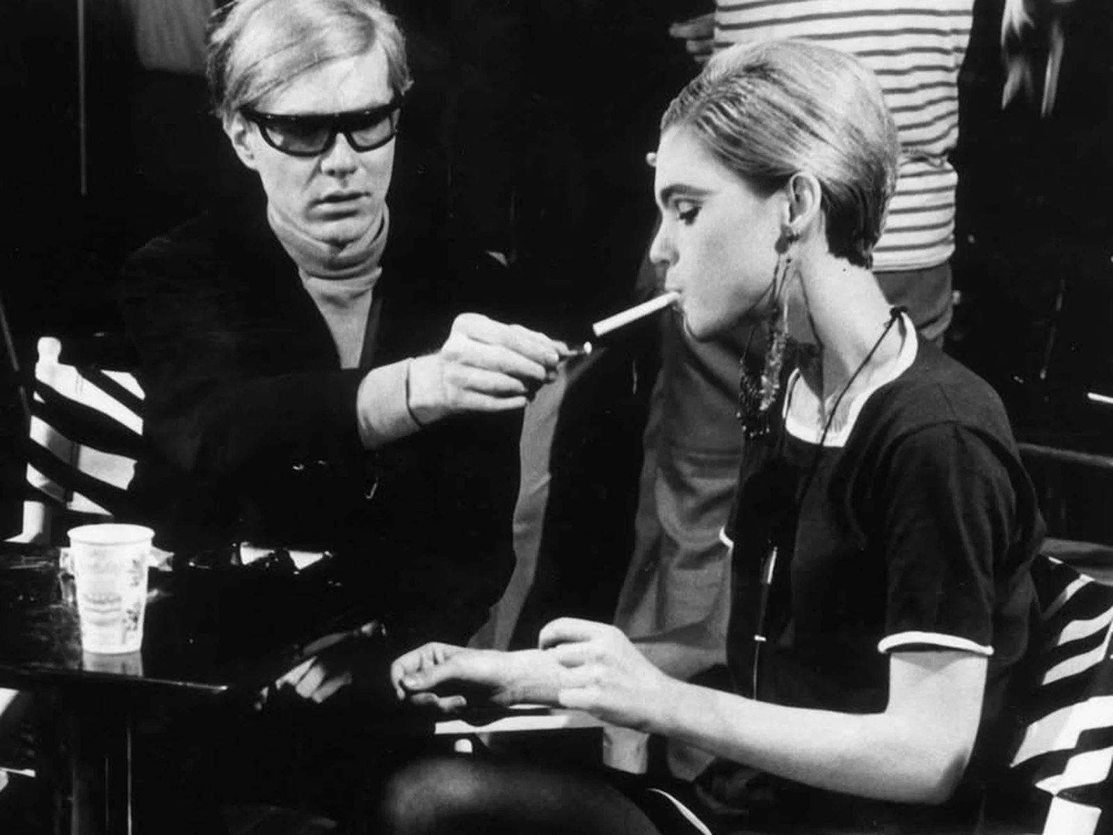 Andy Warhol, lunettes noires, allume la cigarette d'Edie Sedgwick assise sur une chaise de tournage