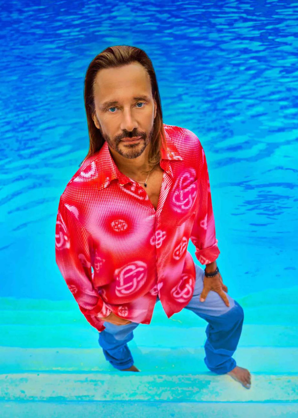 Bob Sinclar en chemise rouge à motifs, les pieds dans l'eau sur les marches de la piscine Molitor