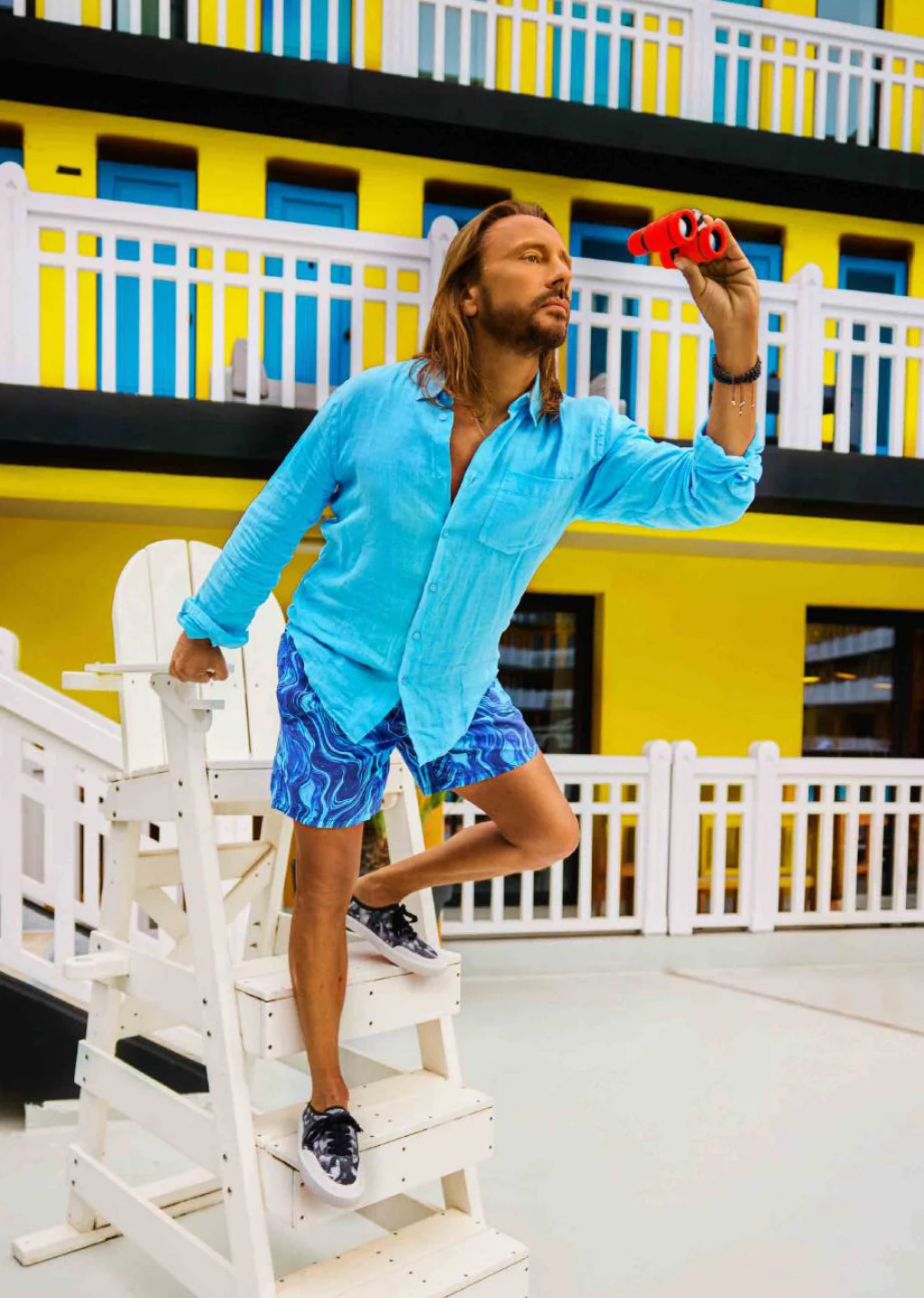 Bob Sinclar en chemise bleue sur une chaise de maître nageur, jumelles rouges à la main, piscine Molitor