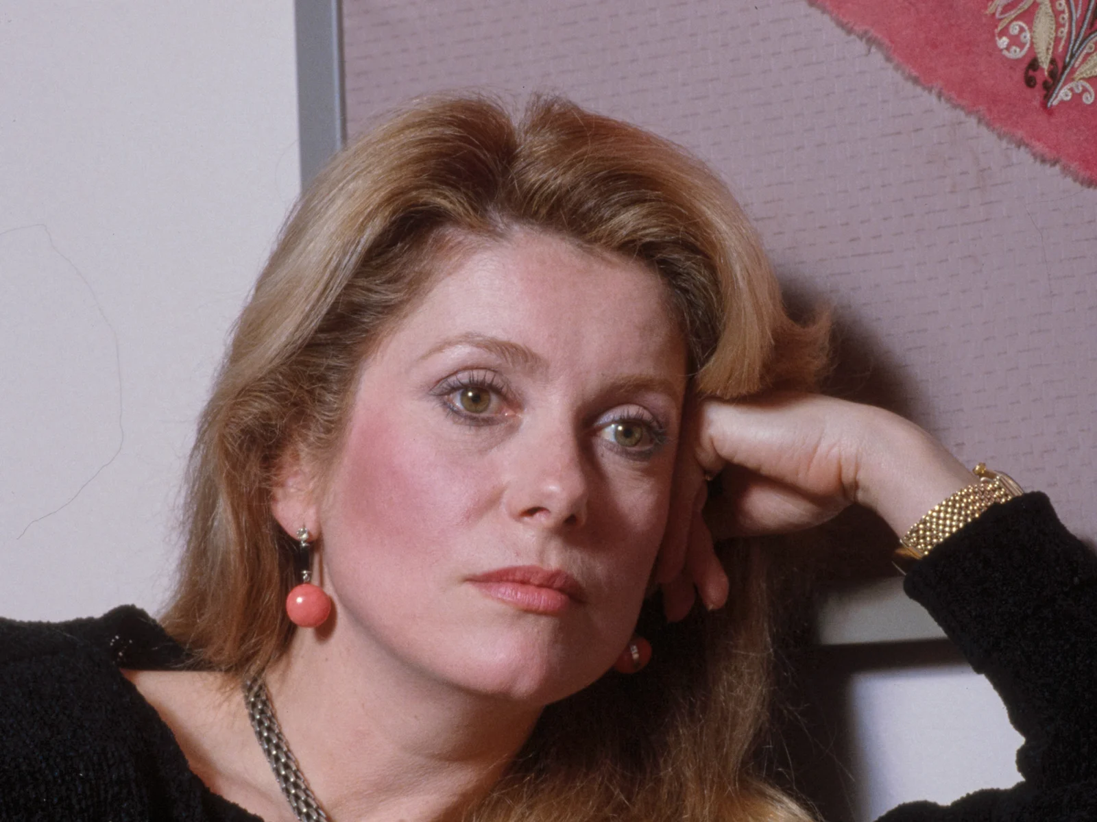 Catherine Deneuve en 1983, pull noir, boucles d'oreilles corail et bracelet doré, la tête posée sur la main, regard pensif