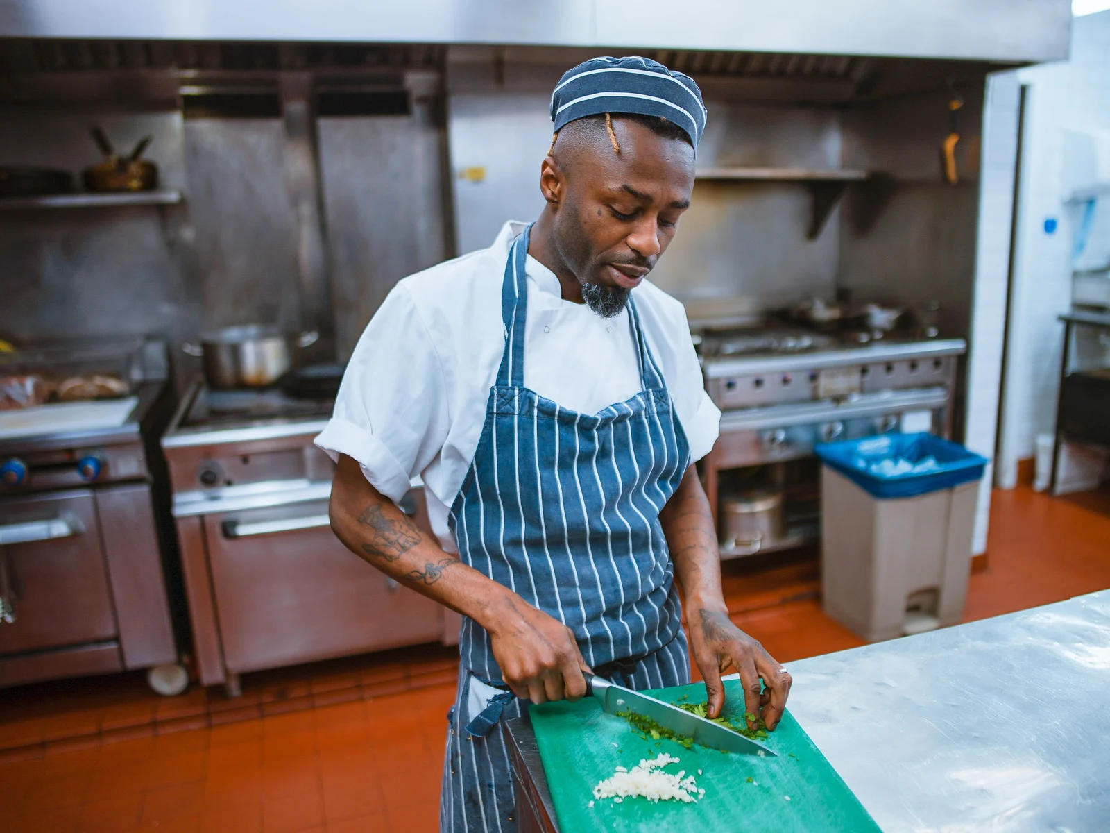Cuisinier en tablier rayé et calot ciselant des herbes sur une planche verte dans une cuisine de restaurant