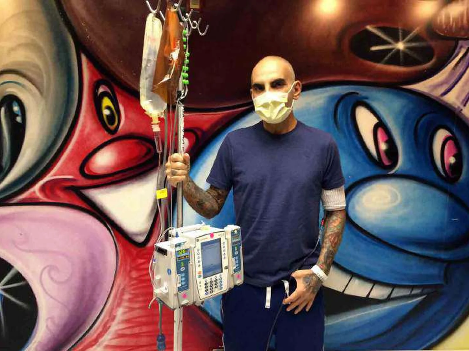 Christian Audigier, masque sur le visage et perfusion à la main, devant une fresque colorée pendant son traitement