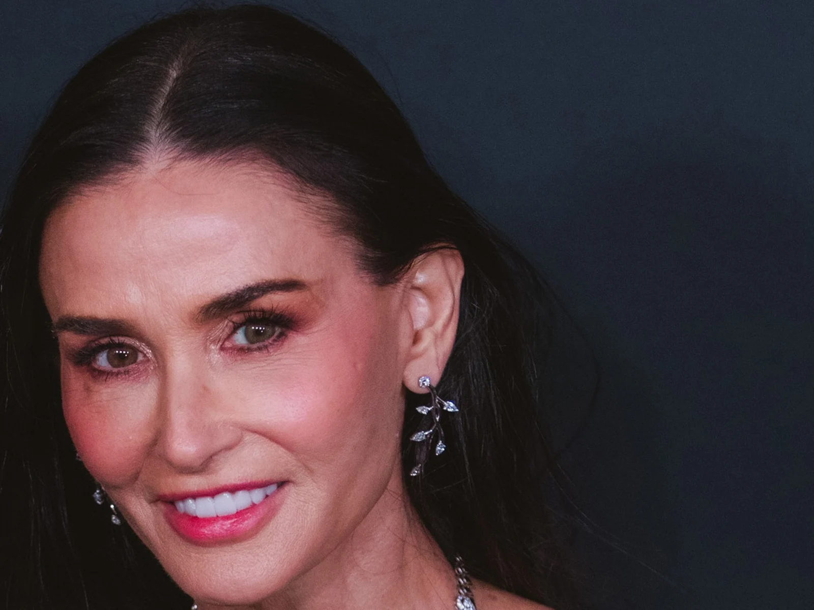 Gros plan sur Demi Moore, cheveux sombres lissés et boucles d'oreilles feuillagées, souriante aux WWD Style Awards 2026