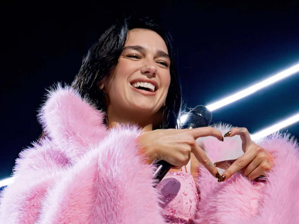 Dua Lipa sourit en manteau de fourrure rose et forme un coeur avec les mains sur scène au Kia Forum, octobre 2025