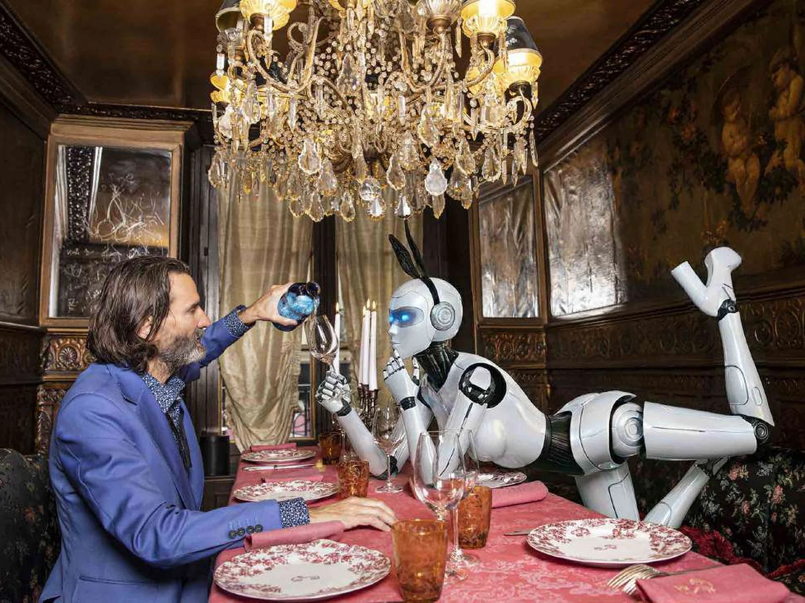 Frédéric Beigbeder en veste bleue, verre à la main, face à un robot humanoïde blanc sur une table sous un lustre