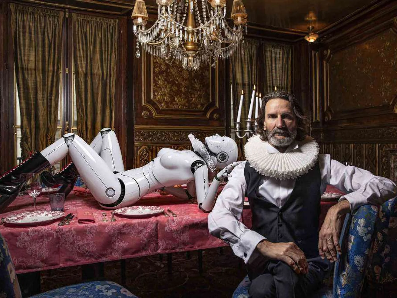 Frédéric Beigbeder en fraise et gilet noir, assis à une table dressée, un robot humanoïde allongé derrière lui