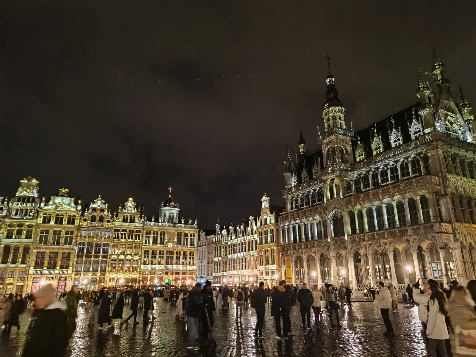 Foule sur les pavés mouillés de la Grand-Place de Bruxelles, la nuit, devant les façades dorées et la Maison du Roi