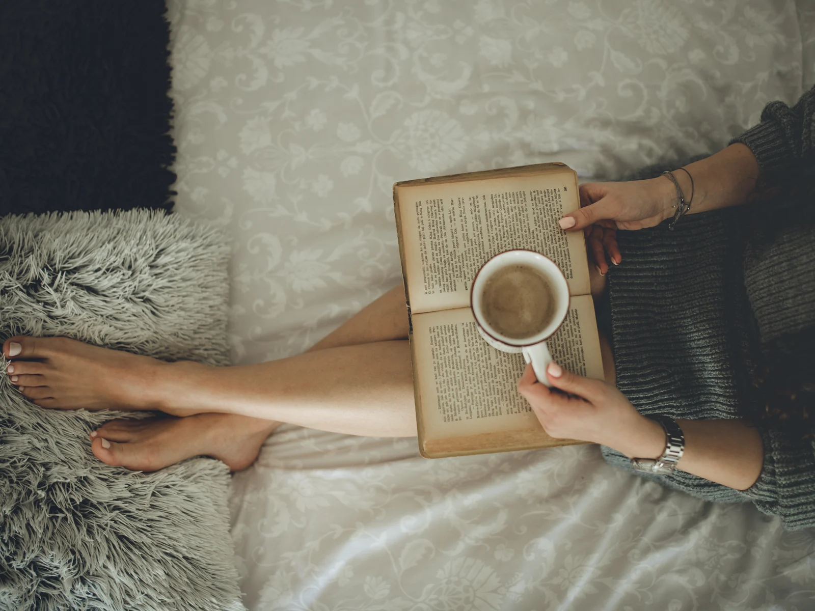 Vue du dessus d'une femme assise sur un lit aux draps brodés, tasse de café posée sur un vieux livre ouvert
