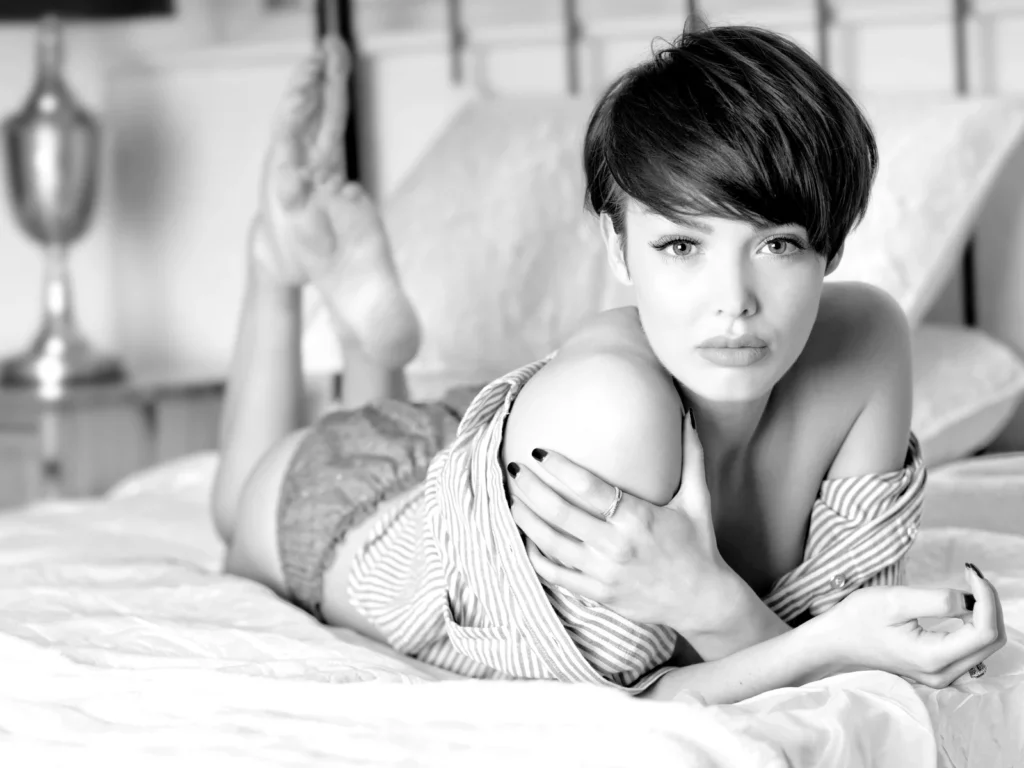 Jeune femme en chemise rayée allongée sur un lit aux draps clairs, portrait noir et blanc pour la grasse matinée
