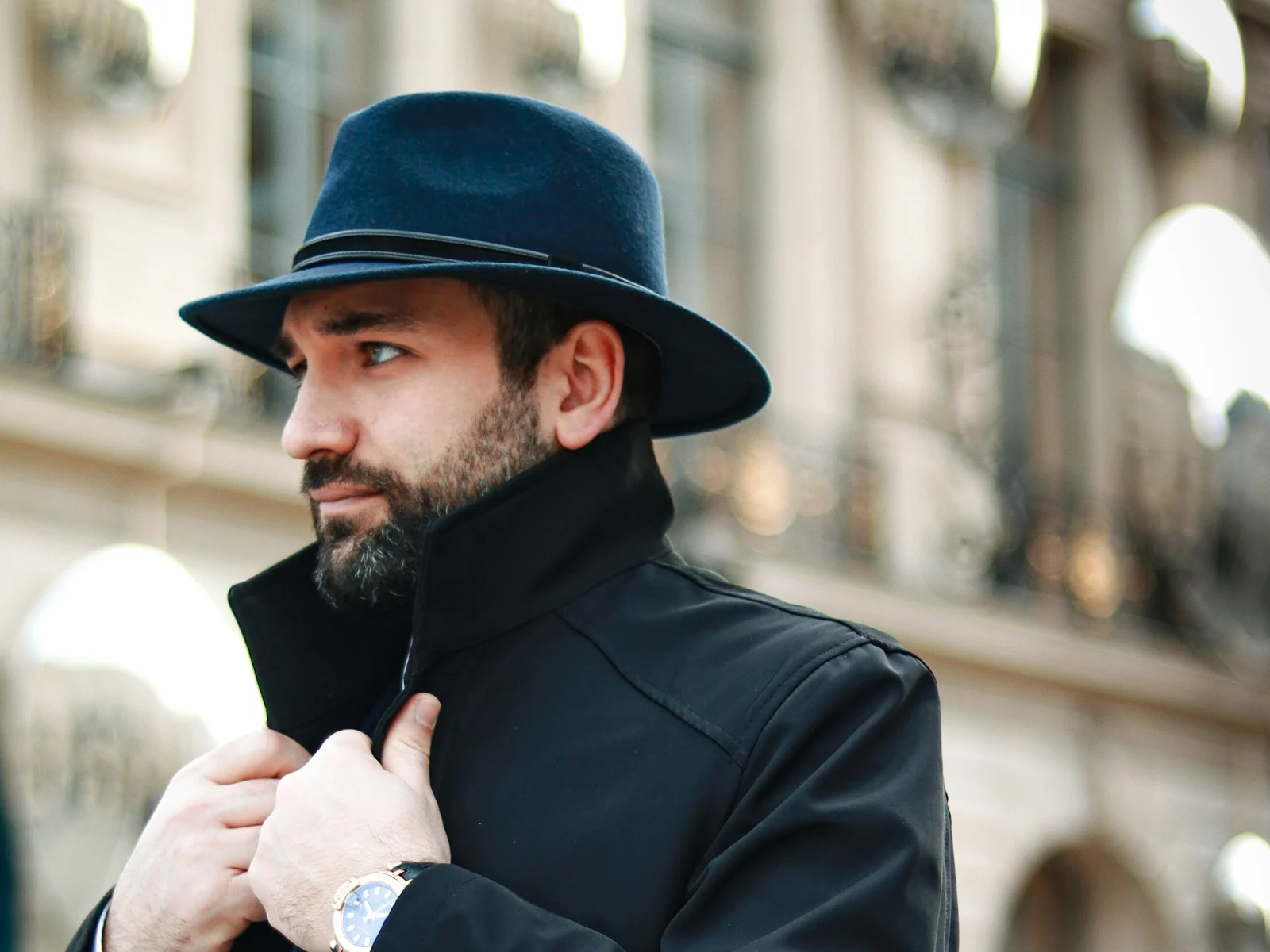 Homme barbu au chapeau de feutre bleu nuit, col relevé d'un long manteau noir, devant une façade haussmannienne à Paris
