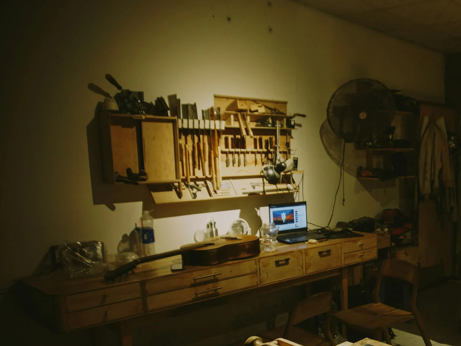 Établi d'atelier, outils accrochés au mur et guitare en réparation dans la pénombre, ambiance du roman Chacun