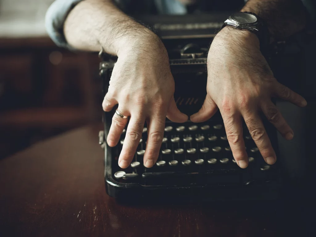 Mains d'homme posées sur les touches d'une machine à écrire ancienne, sur un bureau de bois sombre éclairé en clair-obscur