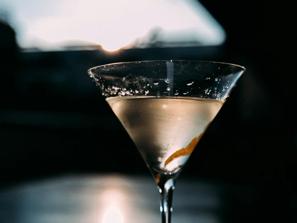 Verre à martini rempli d'un liquide clair et d'un zeste de citron, sur fond sombre et lumière dorée sur le bord