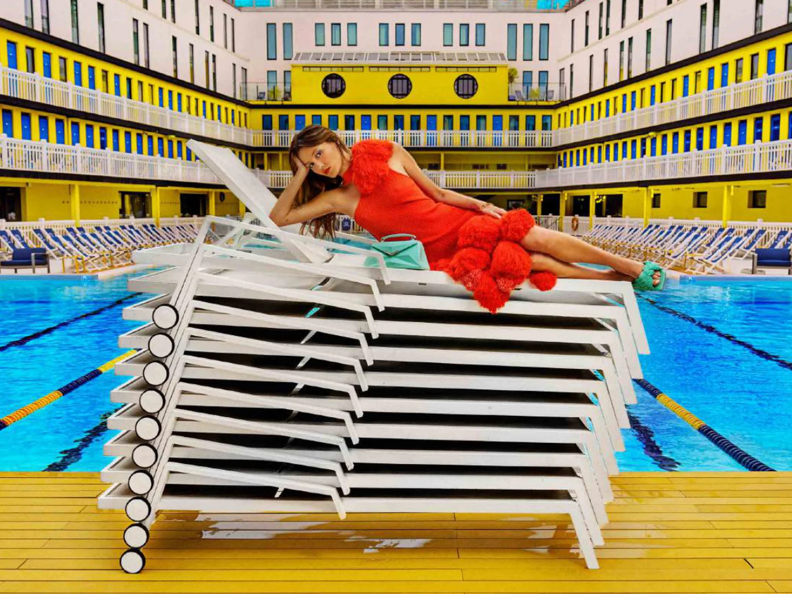 Paloma Le Friant allongée sur une pile de transats devant la piscine Molitor, robe orange à pompons
