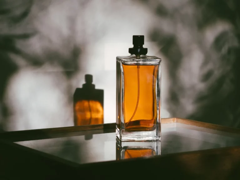 Flacon de parfum ambré sans marque posé sur un plateau miroir, ombres de feuillage en lumière rasante et clair-obscur