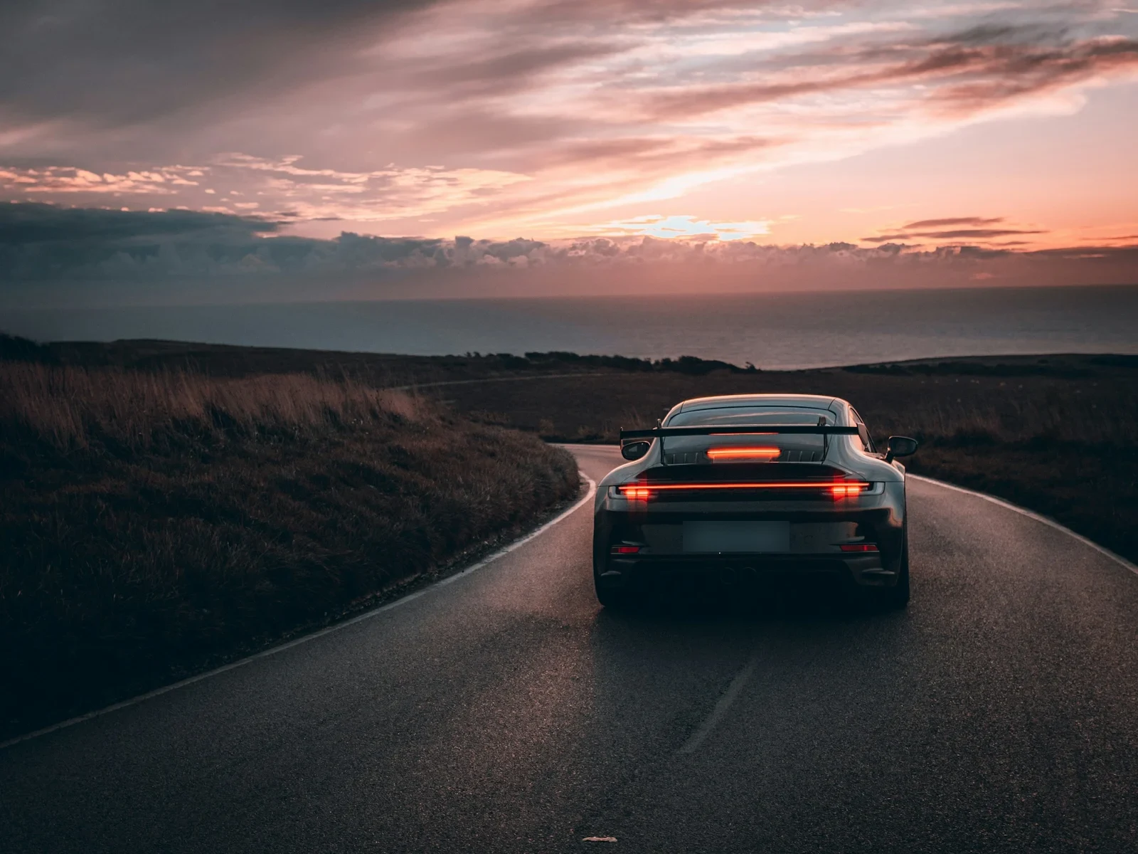 Porsche 911 GT3 vue de dos sur une route côtière sinueuse au crépuscule, feux arrière rouges et ciel rose