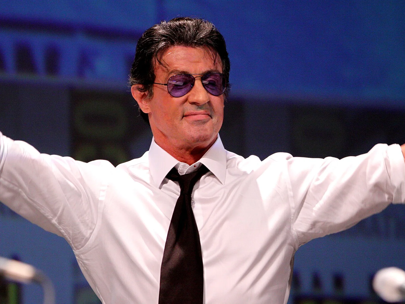 Sylvester Stallone, lunettes aviateur violettes et cravate noire, bras écartés sur scène au Comic-Con de San Diego 2010