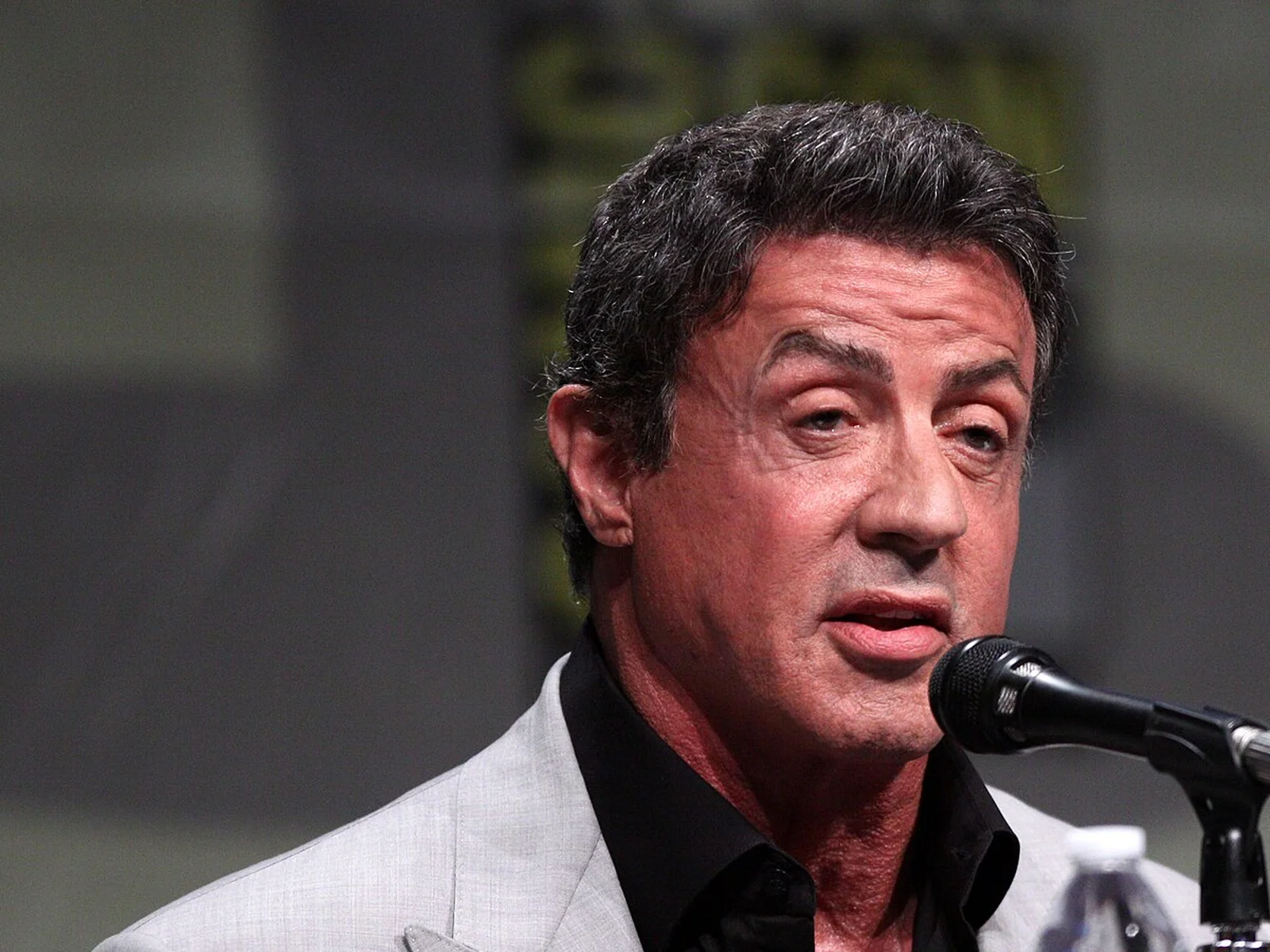 Sylvester Stallone en veste claire et chemise noire prend la parole devant un micro lors du Comic-Con de San Diego 2012