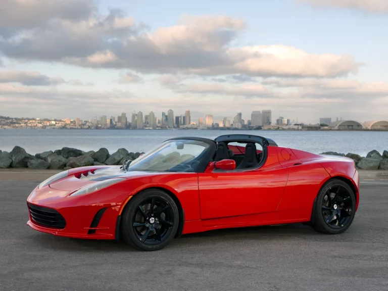Tesla Roadster rouge de première génération, capote ouverte, garée face à la baie et aux gratte-ciel de San Diego